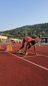 Sprinter im roten Trikot steht im Startblock auf Bahn 5, bereit für den Lauf. Weitere Athleten bereiten sich auf den benachbarten Bahnen vor. Zuschauer und Kampfrichter am Rand der Tartanbahn, im Hintergrund grüne Hügel.