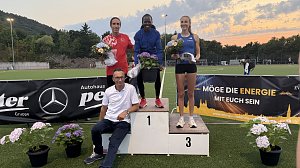 Drei Athlet:innen stehen auf dem Siegerpodest mit Blumen in den Händen, vor ihnen Mercedes-Benz Pkw Verkaufsleiter Jörn Bierwisch mit weißem Shirt. Hinter dem Podest Sponsorenbanner und Abendhimmel.