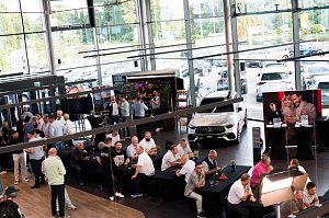 Eine gro&szlig;e Halle mit vielen G&auml;sten, die an schwarzen Tischen sitzen oder an Stehtischen stehen. Ein wei&szlig;er Mercedes ist ausgestellt, dahinter eine Bar und ein Bildschirm mit dem Mercedes-Logo.