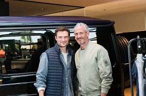 Zwei M&auml;nnermodels stehen nebeneinander vor einem schwarzen Mercedes. Beide l&auml;cheln in die Kamera, einer tr&auml;gt eine helle Bomberjacke, der andere eine dunkle Weste.