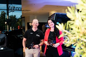 Eine Frau im roten Blazer (Christina Dost) und ein Mann im schwarzen Polohemd (Carsten Dost) stehen nebeneinander und l&auml;cheln. Beide halten Moderationskarten, im Hintergrund ein Mercedes-Gel&auml;ndewagen.