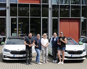 Fünf Personen stehen vor einem modernen Gebäude und posieren mit Champagnerflaschen vor zwei weißen �koda-Fahrzeugen. Auf den Autos sind grüne Elemente und das Logo der Praxisklinik für Zahnheilkunde zu sehen.