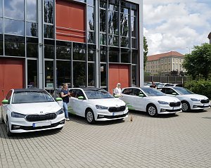 Vier weiße �koda-Fahrzeuge mit grünen Akzenten stehen nebeneinander vor einem modernen Gebäude. Vor den Autos stehen mehrere Personen mit Flaschen in der Hand, wie bei einer feierlichen Fahrzeugübergabe.