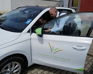 Ein Mann im blauen Sakko zeigt einer Frau auf dem Fahrersitz eines weißen �koda-Fahrzeugs etwas auf einem Tablet. Auf der Autotür ist das Logo der Praxisklinik für Zahnheilkunde zu sehen.