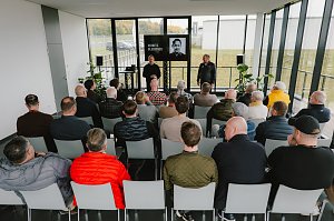 Eine Gruppe von Menschen sitzt in einem modernen, hellen Raum w&auml;hrend einer Pr&auml;sentation. Im Vordergrund sieht man mehrere M&auml;nner, die auf St&uuml;hlen sitzen und auf eine B&uuml;hne blicken. Auf der B&uuml;hne stehen zwei Personen: Ein Mann tr&auml;gt einen schwarzen Pullover und spricht, w&auml;hrend der andere in einem schwarzen Outfit steht. Auf einem Bildschirm im Hintergrund wird ein Portr&auml;t von Moritz Bleibtreu gezeigt. Der Raum hat gro&szlig;e Fenster, durch die man die Natur drau&szlig;en sieht. Pflanzen stehen in den Ecken des Raumes.
