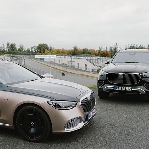 Zwei Luxusautos von Mercedes-Benz stehen nebeneinander auf einer Rennstrecke. Auf der linken Seite ist ein silberner Mercedes-Maybach S-Class zu sehen, der eine gl&auml;nzende schwarze Motorhaube und schwarze R&auml;der hat. Rechts daneben steht ein schwarzer Mercedes-Maybach GLS, ein gro&szlig;er SUV mit auff&auml;lligem K&uuml;hlergrill und silbernen Akzenten. Im Hintergrund erstreckt sich die Rennstrecke, und B&auml;ume mit herbstlichen Farben sind am Horizont sichtbar.