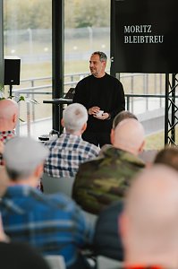 Ein Mann in schwarzem Pullover h&auml;lt eine Tasse in der Hand und spricht zu einem Publikum. Auf dem Bildschirm hinter ihm ist der Name 'Moritz Bleibtreu' zu sehen. Die Zuschauer im Vordergrund sind nur von hinten zu sehen.