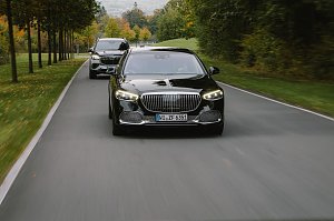 Ein schwarzer Mercedes-Maybach S-Class f&auml;hrt auf einer Landstra&szlig;e, gefolgt von einem Mercedes-Maybach SUV. Beide Fahrzeuge sind von vorne zu sehen, mit dem S-Class im Vordergrund und dem SUV dahinter. Die Stra&szlig;e wird von B&auml;umen ges&auml;umt, und die Farben des Herbstes sind im Hintergrund sichtbar.