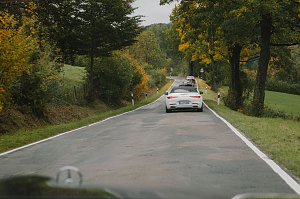 Eine Gruppe von Fahrzeugen f&auml;hrt auf einer kurvigen Landstra&szlig;e, die von B&auml;umen ges&auml;umt ist. Im Vordergrund ist ein wei&szlig;er Mercedes-Benz Cabrio zu sehen, gefolgt von weiteren Fahrzeugen. Die B&auml;ume zeigen herbstliche Farben, und die Stra&szlig;e ist von gr&uuml;nen Wiesen und einem h&uuml;geligen Hintergrund umgeben.