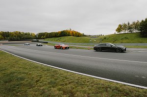 Mehrere Autos fahren auf einer Rennstrecke. Im Vordergrund ist ein schwarzer Mercedes-AMG und ein oranger Mercedes-AMG GT zu sehen. Weitere Fahrzeuge sind in der Ferne sichtbar, w&auml;hrend die Strecke von B&auml;umen ges&auml;umt ist, die herbstliche Farben zeigen. Der Himmel ist bew&ouml;lkt, und die Strecke f&uuml;hrt in eine Kurve.