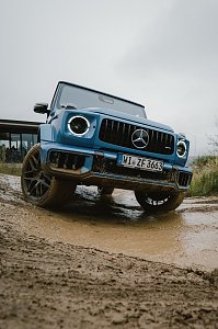 Ein blauer Mercedes-Benz G-Class f&auml;hrt auf einem matschigen Gel&auml;nde. Der Blickwinkel zeigt das Fahrzeug aus der N&auml;he, mit dem Fokus auf die Frontpartie und die markante K&uuml;hlergrill. Der Unterboden und die R&auml;der sind mit Schlamm bedeckt, was die Gel&auml;ndetauglichkeit des Fahrzeugs betont. Im Hintergrund ist ein Geb&auml;ude unscharf zu sehen.