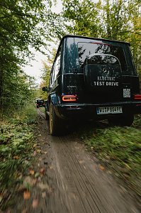 Ein Mercedes-Benz G-Class, Modell G 580, f&auml;hrt auf einem unbefestigten Waldweg. Das Fahrzeug ist mit dem Aufkleber 'ELECTRIC TEST DRIVE' auf der Heckklappe versehen. Die Reifen sind mit Schlamm bedeckt, und im Hintergrund ist ein weiteres Fahrzeug zu sehen. Der Wald mit gr&uuml;nen B&auml;umen umgibt den Weg.