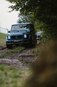 Ein blauer Mercedes-Benz G-Class f&auml;hrt auf einem unbefestigten, felsigen Weg im Wald. Der Blick ist von der Seite, sodass die Vorderseite des Fahrzeugs und die charakteristischen Scheinwerfer gut sichtbar sind. Im Hintergrund ist ein weiteres Fahrzeug zu sehen. Der Weg ist von Gras und B&auml;umen ges&auml;umt.