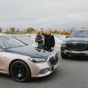 Zwei M&auml;nner stehen l&auml;chelnd nebeneinander zwischen zwei Mercedes-Maybach Fahrzeugen auf dem Gel&auml;nde der Mercedes-Benz Driving Experience am Bilster Berg. Im Hintergrund ist die Rennstrecke mit herbstlicher Landschaft zu sehen.