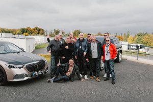 Eine Gruppe von M&auml;nnern steht lachend zwischen zwei Mercedes-Maybach Fahrzeugen auf dem Gel&auml;nde der Mercedes-Benz Driving Experience am Bilster Berg. Einige tragen bunte Jacken, und einer sitzt entspannt auf dem Boden. Im Hintergrund sind Geb&auml;ude der Rennstrecke und herbstlich gef&auml;rbte B&auml;ume zu sehen.