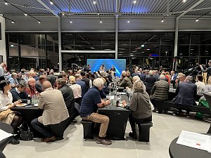 Frontale Ansicht der B&uuml;hne mit CDU-Hintergrund. Zwei Personen sprechen, w&auml;hrend das Publikum an schwarzen Tischen zuh&ouml;rt. Der Raum ist hell beleuchtet und modern eingerichtet.