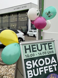 Werbeschild mit Text Heute 10:00�14:00 Uhr Skoda Buffet, dekoriert mit bunten Luftballons vor einem Autohaus.