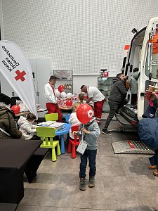 Kinder basteln an einem Tisch mit Rotkreuz-Ballons, daneben Sanitäter und ein offener Rettungswagen im Autohaus.
