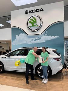 Mann und junge Frau in grünen Shirts stehen vor einem weißen �koda im Autohaus. Die junge Frau hält einen silbernen Behälter, der Mann greift hinein und zieht einen Umschlag.