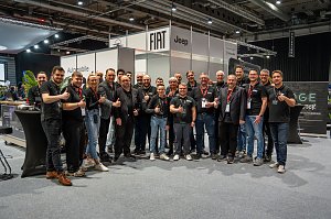 Gruppe von Mitarbeitenden posiert lächelnd mit Daumen hoch an einem Messestand in einer Messehalle.