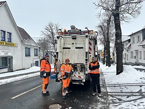 Drei Mitarbeiter der Müllabfuhr stehen hinter einem Müllfahrzeug auf einer verschneiten Straße.