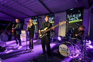 Eine Live-Band sorgt für Stimmung: Sängerin, Gitarrist, Bassist und Schlagzeuger stehen auf einer Bühne mit moderner Lichttechnik und performen vor den Gästen. Im Hintergrund mehrere Displays mit der Aufschrift Luxus Loft � Die Party WG.