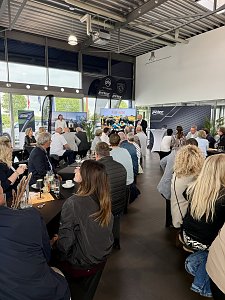 Szene im Peugeot-Autohaus: viele Personen sitzen an mehreren Tischen, während davor eine Person ins Mikrofon spricht.