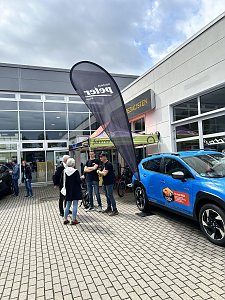 Hof vor Autohaus mit Beachflag, blauem Fahrzeug und einigen Personen, die sich unterhalten.


