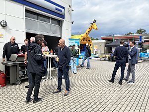 Hof vor Autohaus mit Grill, H&uuml;pfburg im Hintergrund und einigen Personen, die sich unterhalten.

