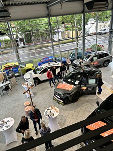 Autohaus von innen mit Holzwegweiser und Sitzs&auml;cken in Holzstumpf-Optik und mehreren Personen, die sich die Autos anschauen.


