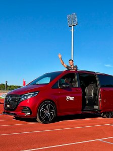 Ein Top-Athlet winkt gut gelaunt aus dem ge&ouml;ffneten Schiebedach eines roten Mercedes-Benz EQV des Autohauses Peter w&auml;hrend der Einfahrt auf der Stadionlaufbahn.