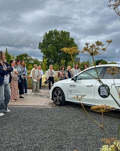 Eine größere Gruppe von Frauen, die sich im Freien unter bewölktem Himmel versammelt haben und aufmerksam einen weißen Mercedes-Benz CLA betrachten.