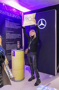 Eine Frau in dunkler Kleidung, die freudig ein Kissen mit der Aufschrift She�s Mercedes vor einer Mercedes-Eventwand in die Höhe hält.
