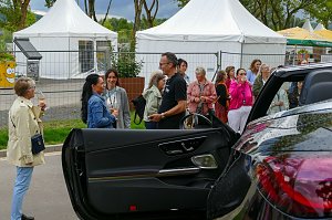 Eine Menschengruppe im Freien, die sich im Hintergrund unterhält, während im Vordergrund die geöffnete Fahrertür eines dunklen Mercedes-Cabriolets zu sehen ist.
