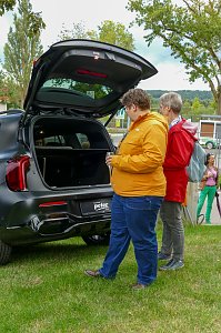 Zwei Frauen in farbigen Jacken, die im Freien auf einer Wiese stehen und den geöffneten Kofferraum eines schwarzen Mercedes-Fahrzeugs betrachten.