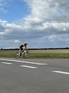 Radstrecke beim Triathlon. Ein Athlet im grünen Einteiler und mit weißem Helm fährt auf einem hellen Rennrad vorbei. Dahinter erstreckt sich eine weite Feldlandschaft unter einem leicht bewölkten Himmel.