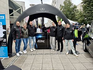 Man sieht das Team vom Autohaus Peter vor einem schwarzen, aufblasbaren Pavillon mit Mercedes-Benz Aufdruck. In der Mitte steht ein schwarzer Counter, mit Logo-Aufdruck der Autohaus Peter Gruppe in Weiß.