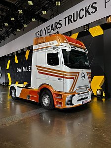 Weiß-orangefarbener Mercedes Lkw von vorne rechts auf einer Messe. Am Fahrerhaus kleben gold-rote Streifen. Im Hintergrund hängt ein Banner mit der Aufschrift 130 YEARS TRUCKS. Der Lkw steht auf einem glänzenden, spiegelnden Messeboden.