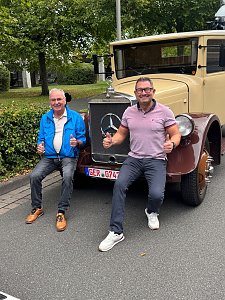 Zwei lachende Männer sitzen auf den Kotflügeln eines beigefarbenen Oldtimer-Lkws mit braunen Radläufen. Beide zeigen den Daumen nach oben. Der Mann links trägt eine blaue Jacke, der rechte ein rosa Poloshirt. Im Hintergrund stehen grüne Bäume.