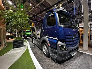 Dunkelblauer Mercedes-Benz Lkw von vorne rechts auf einem Messestand. Im Hintergrund links steht ein grünes Nutzfahrzeug neben einem großen, echten Baum im Pflanzkübel. Der Boden hat weiße Linien und grüne Kunstrasen-Flächen.