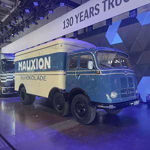 Historischer Mercedes-Benz Lkw auf einer Messe. Blaues Fahrerhaus und cremeweißer Kofferaufbau mit der Aufschrift MAUXION SCHOKOLADE. Drei Achsen mit schwarzen Felgen. Im Hintergrund hängt ein Banner mit 130 YEARS TRUCKS.