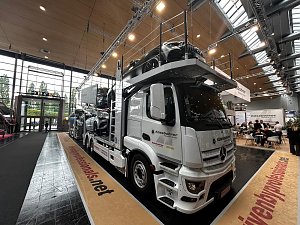 Weißer Mercedes-Benz Autotransporter schräg von vorne in einer großen Messehalle. Mehrere Pkws sind auf den Rampen über dem Fahrerhaus und auf dem Anhänger geladen. Der Lkw steht auf einer beigen Ausstellungsfläche mit roter Schrift.