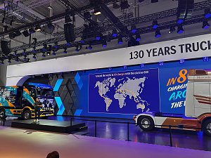 Große Messebühne mit zwei Mercedes-Benz Lkws. Links ein bunt beklebter Lkw auf einem Podest, rechts das Heck einer weiß-orangen Zugmaschine. Im Hintergrund leuchtet eine blaue LED-Wand mit einer Weltkarte unter dem Banner 130 YEARS TRUCKS.