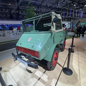 Grüner historischer Mercedes-Benz Unimog schräg von vorne auf einem hellen Holzboden auf einer Messe. Er hat auffällige, rote Felgen, eine eckige Front und ein beiges Stoffverdeck. Im Hintergrund stehen Messebesucher. Schwarze Absperrstangen davor.