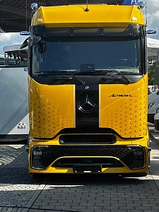 Gelber Mercedes-Benz Lkw direkt von vorne fotografiert. In der Mitte der Front verläuft ein breiter, schwarzer Längsstreifen mit dem Markenlogo. Das Fahrzeug steht auf einem gepflasterten Außenbereich.