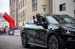 Man sieht das F&uuml;hrungsfahrzeug, den vollelektrischen GLC. Aus dem Beifahrerfenster heraus fotografiert eine Frau. Das Auto steht. Im Hintergrund sind die L&auml;ufer und L&auml;uferinnen zu sehen.