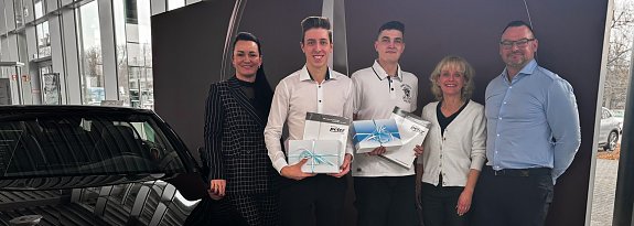Christina Dost, Gesch&auml;ftsf&uuml;hrerin, steht links neben Paul W. (zweiter von links), der nach verk&uuml;rzter Ausbildung als Assistent der Gesch&auml;ftsleitung startet, und Domenik B. (Mitte), der das Team k&uuml;nftig als Karosseriebauer verst&auml;rkt. Rechts daneben stehen Silke Lang, Ausbildungsleiterin, und Enrico Dost, Serviceleiter. Paul W. und Domenik B. halten jeweils eine Geschenkbox mit hellblauer Schleife in den H&auml;nden. Die Gruppe steht im Autohaus vor einem schwarzen Auto und einer Wanddekoration und l&auml;chelt in die Kamera.
