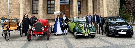 Gruppenfoto mit Fahrzeugen verschiedener Epochen: Vor einem historischen Geb&auml;ude stehen vier Autos � von einer Kutsche mit Holzaufbau &uuml;ber einen roten Oldtimer, einen gr&uuml;nen Oldtimer-Cabrio bis zu einem modernen schwarzen Mercedes. Davor posieren Personen in historischer Kleidung und moderner Garderobe.