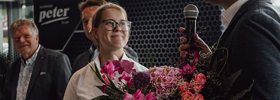Lilly Hecker, frisch ausgebildete Automobilkauffrau und Jahrgangsbeste, steht l&auml;chelnd mit einem Blumenstrau&szlig; neben Gesch&auml;ftsf&uuml;hrer Andreas Peter, der in ein Mikrofon spricht. Im Hintergrund sind weitere Personen und das Logo der Autohaus-Peter-Gruppe zu sehen.