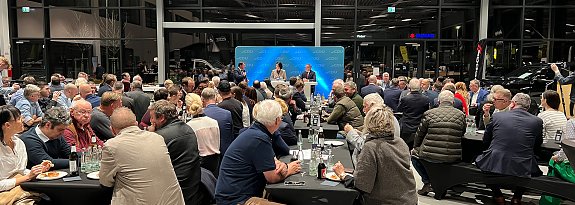 Eine gro&szlig;e Gruppe von Menschen sitzt an langen, schwarz gedeckten Tischen in einer modernen Autohaus-Halle. Vorne steht eine B&uuml;hne mit CDU-Logo, auf der zwei Personen sprechen. Im Hintergrund sind Glasfassaden und Autos sichtbar.